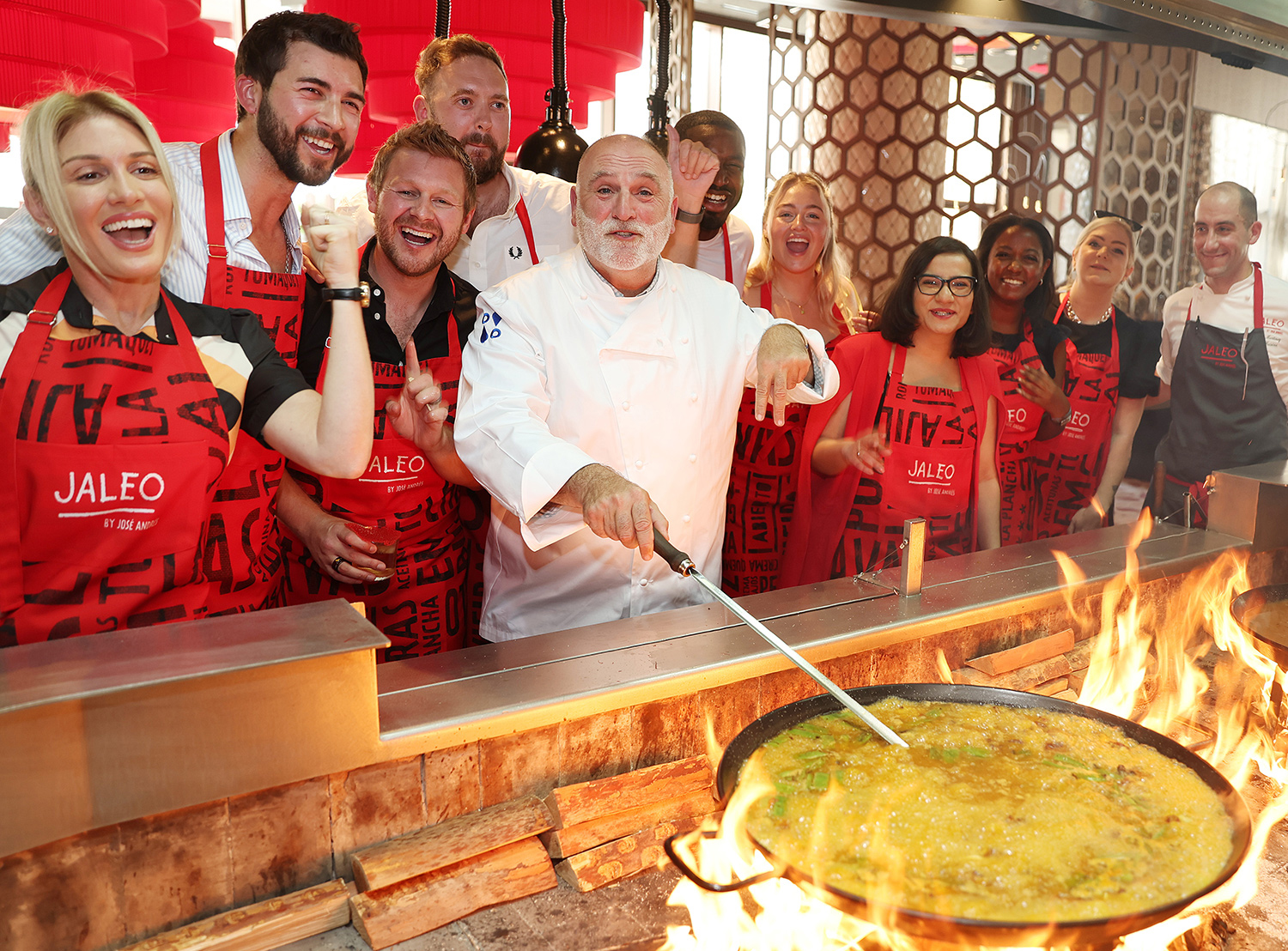 José Andrés stirs paella over a flame as several people in red aprons surround him and smile.