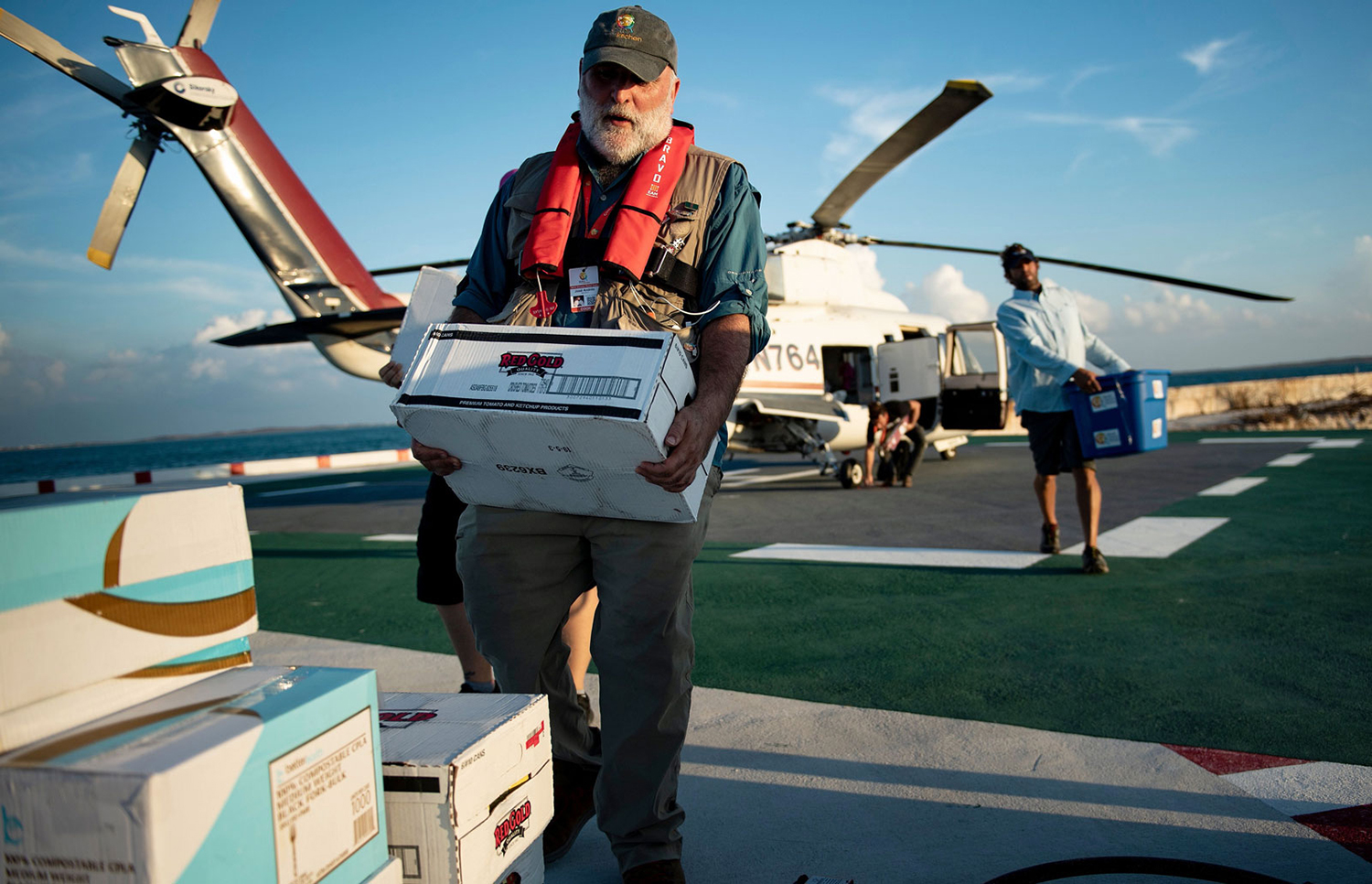 José Andrés walks away from a helicopter while carrying a box toward a pile of similar boxes.