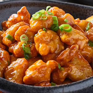 Orange chicken is in a bowl and next to a bowl of white rice and some cut up oranges.