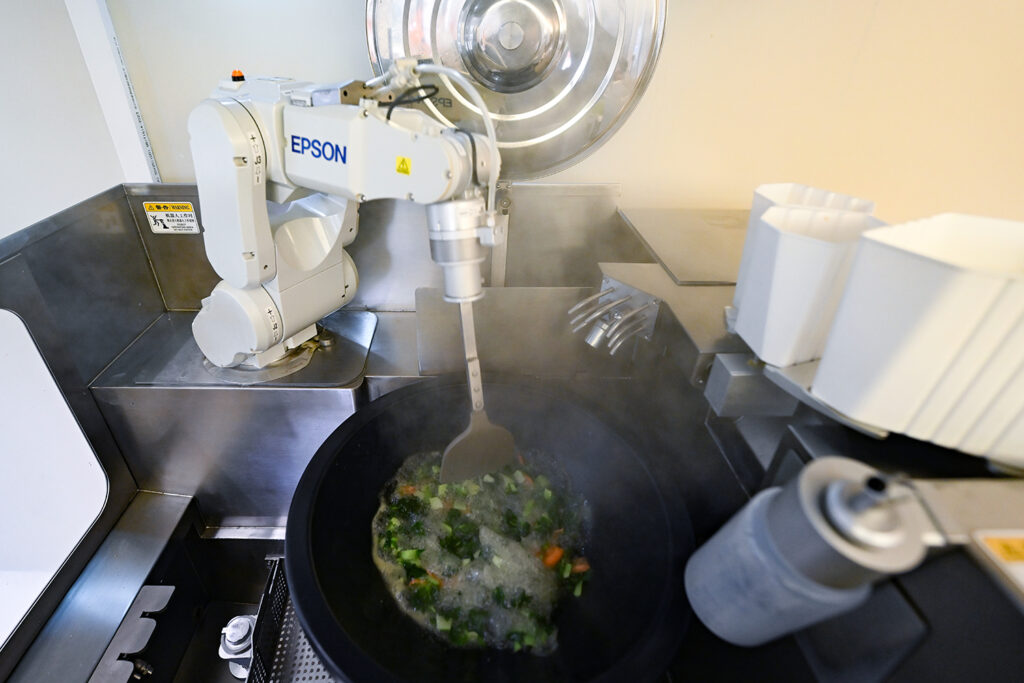A robot arm holds a spatula to stir vegetables that are cooking in a wok.