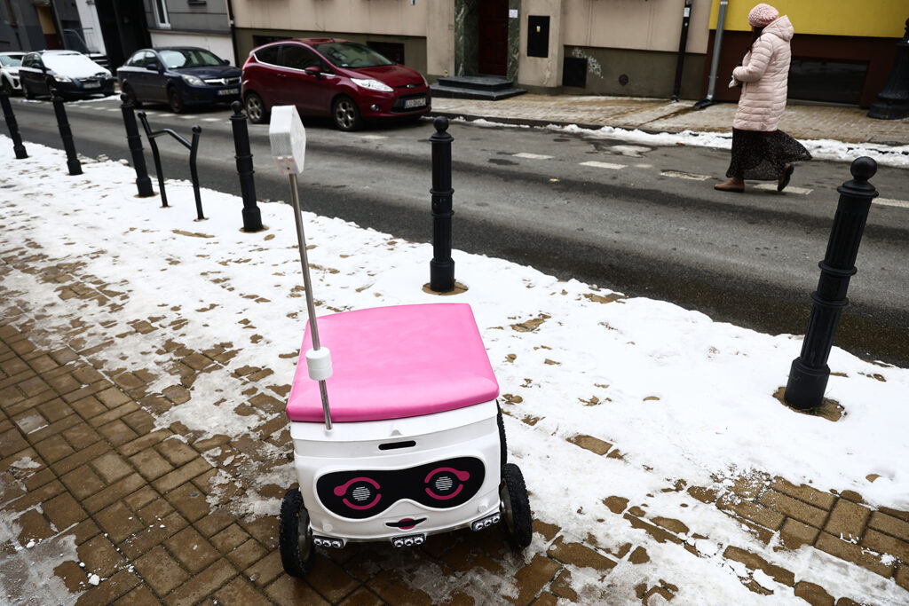 A white cube-shaped robot with a pink lid is on a snowy city sidewalk.