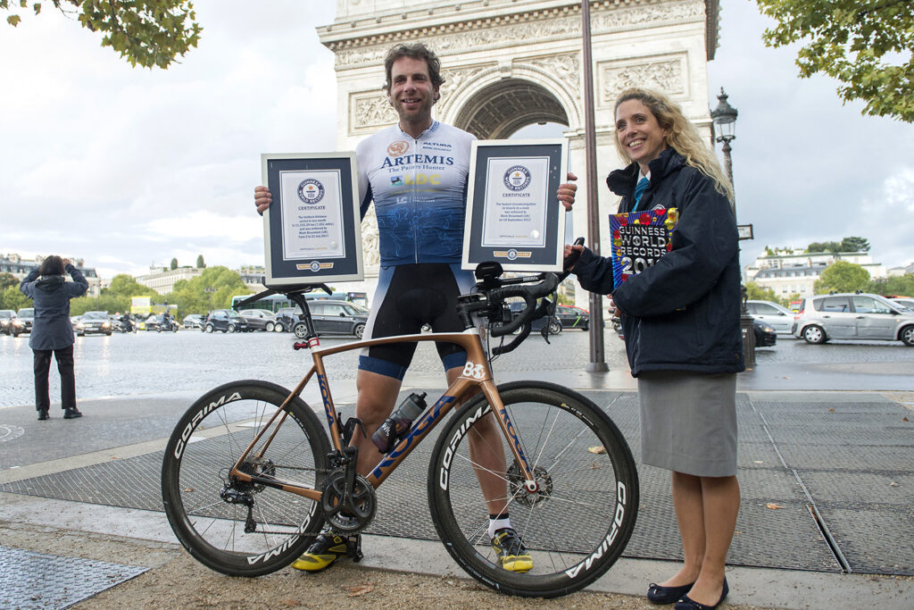 Mark Beaumont and a woman pose with his bike in front of the Arc de Triomphe in Paris holding two certificates from Guinness World Records.