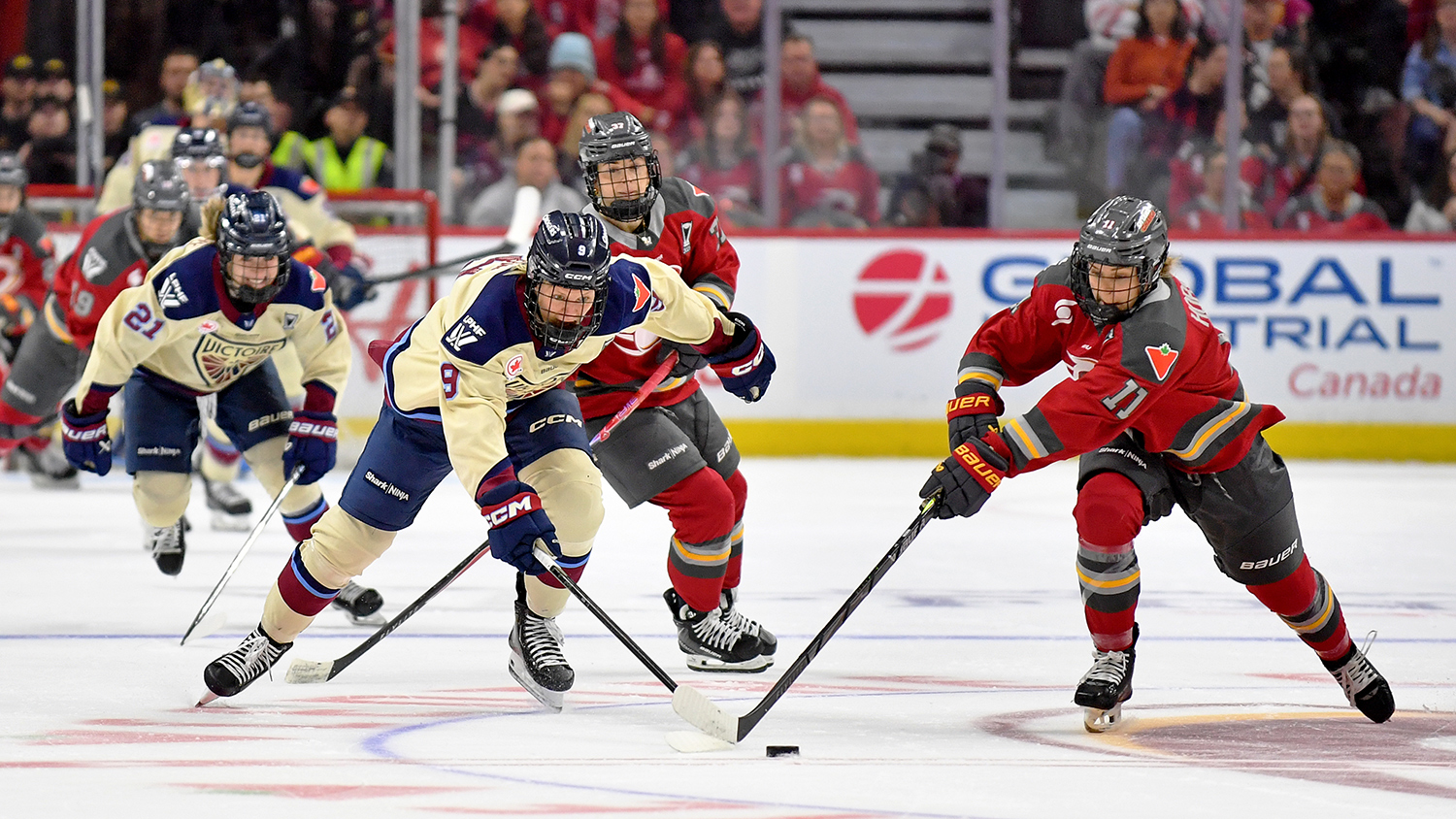 Two hockey players use their sticks to try to gain control of a puck in front of two other players and a crowd of spectators.