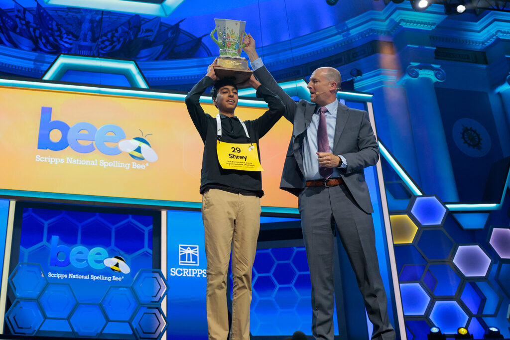 A teenager stands on a stage holding a trophy over his head next to a sign reading Scripps National Spelling Bee, and a man stands next to him.