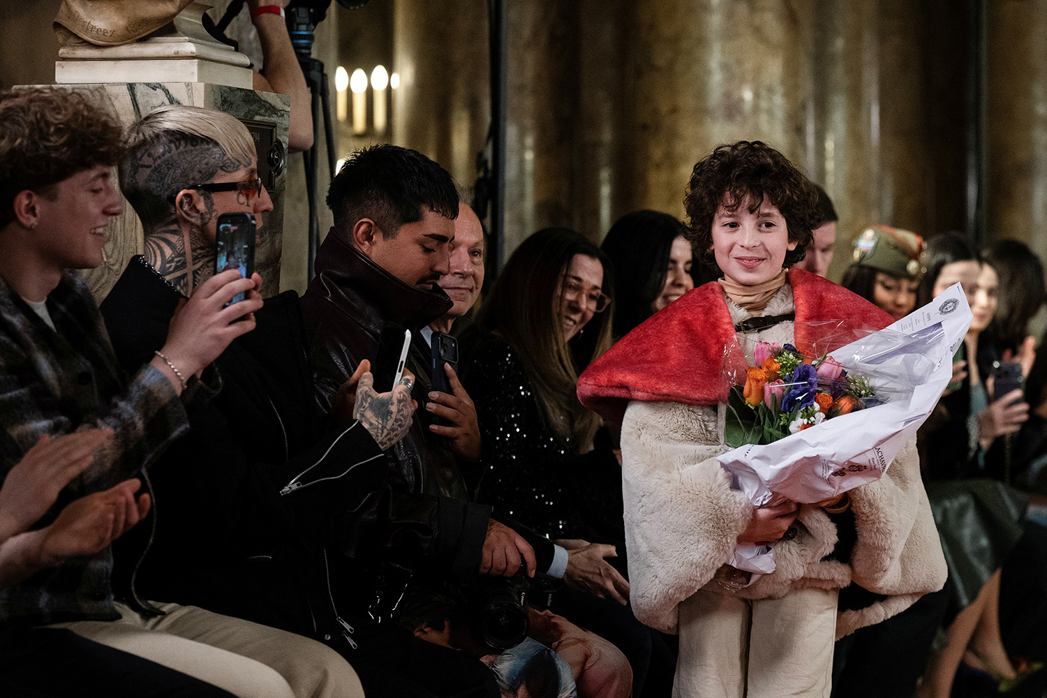 Max Alexander stands on a runway holding flowers as seated audience members smile and some take his photo.