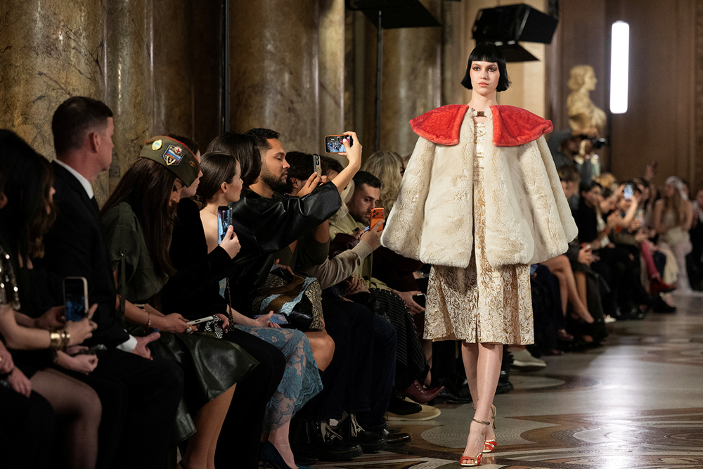A model walks down the runway wearing an off-white cape with a red collar.