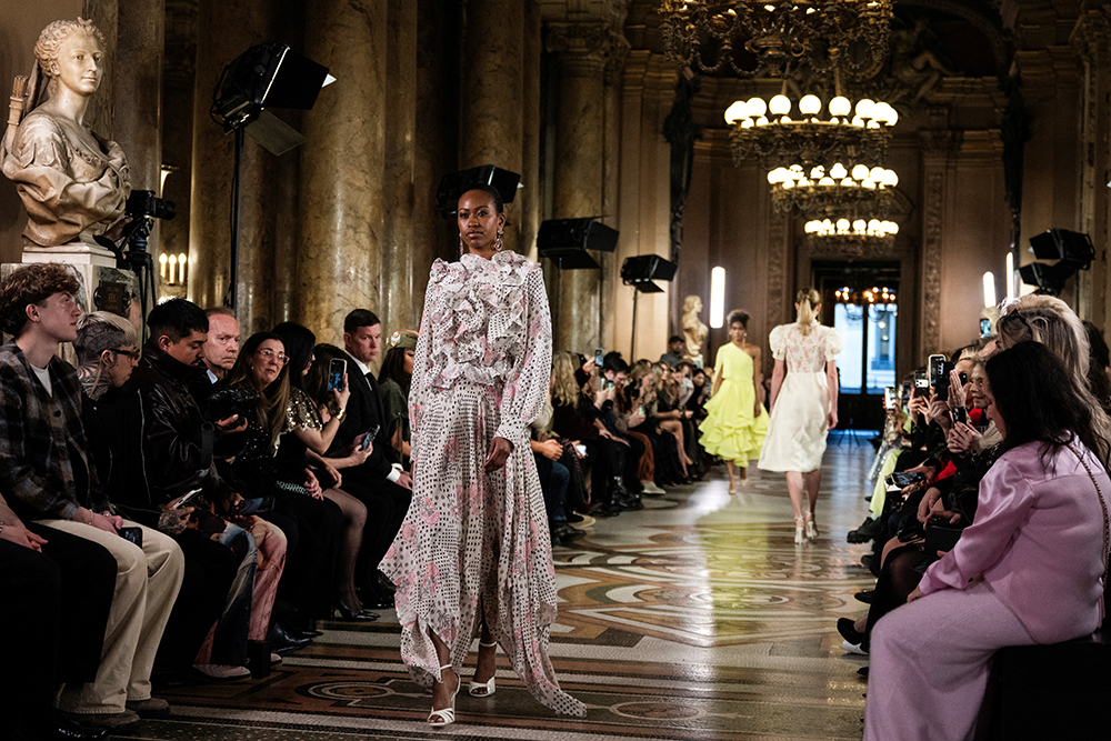 Three models walk the runway in dresses as people are seated and watching along both sides.
