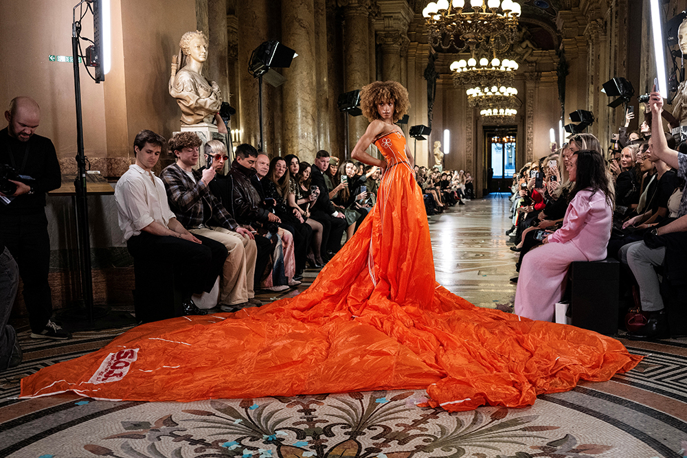 A model stands on a runway wearing a red strapless dress with a large train.