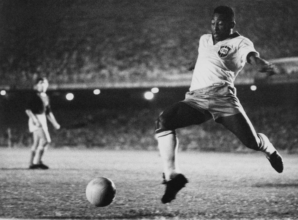 Young Pelé is on a soccer field poised to kick the ball as spectators and a member of the opposing team are seen in the background.