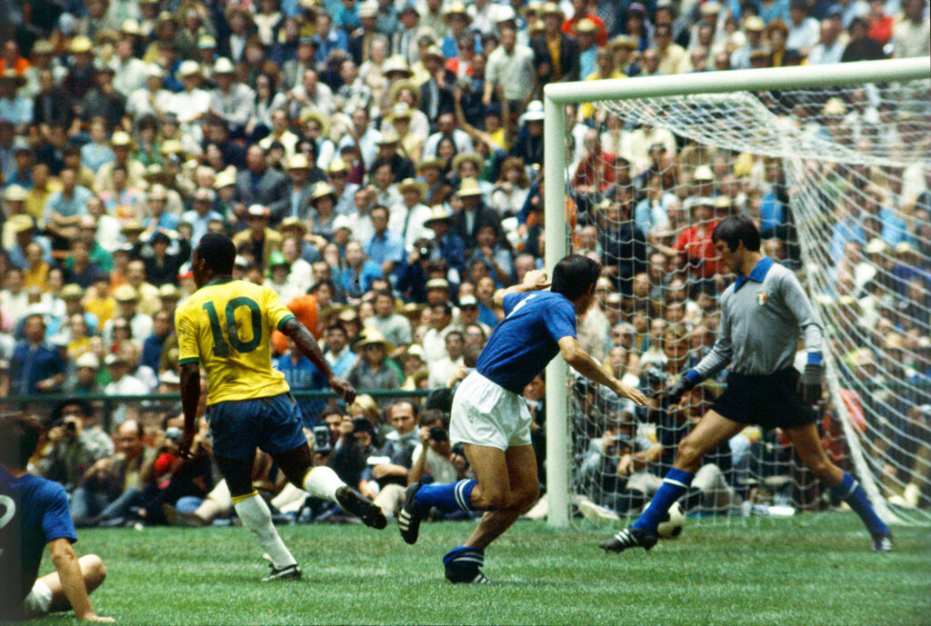 Pelé’s back is to the camera as he runs beyond a goal containing a goalie and a soccer ball.