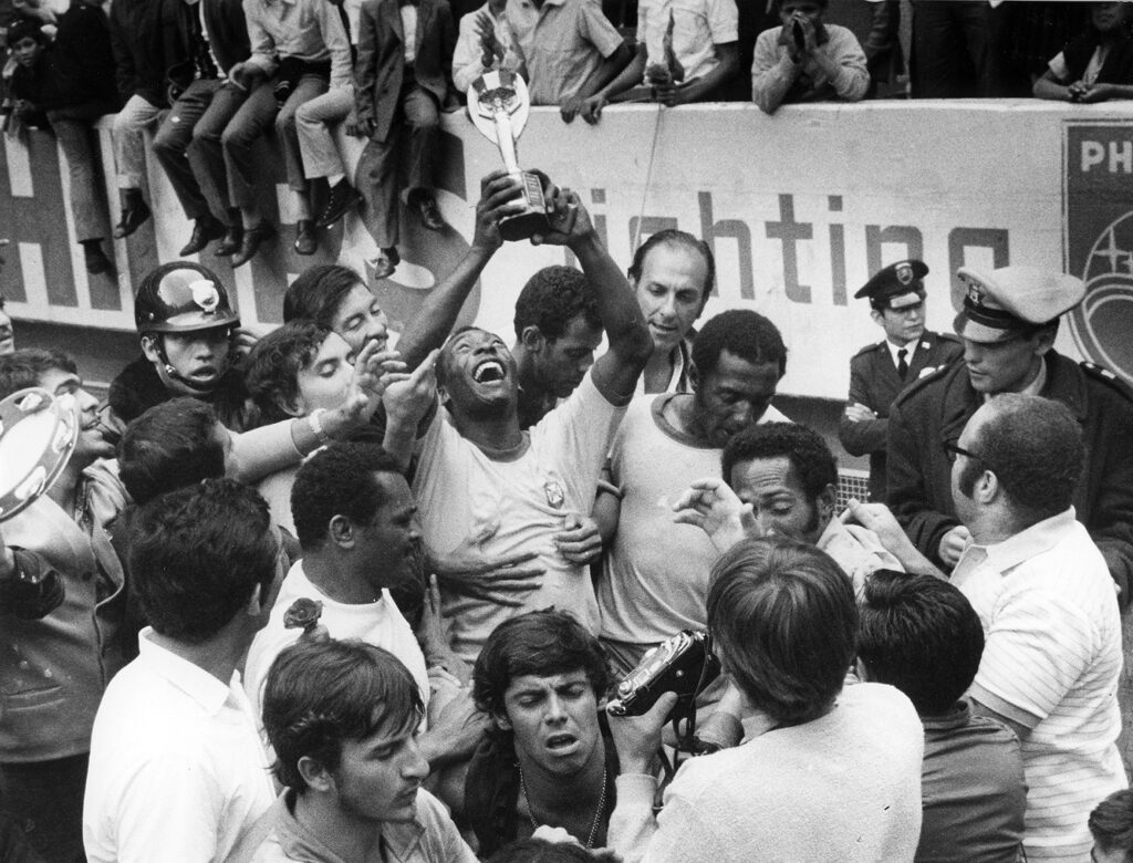 Pelé smiles and holds up a trophy and is surrounded by teammates, police officers, and others.