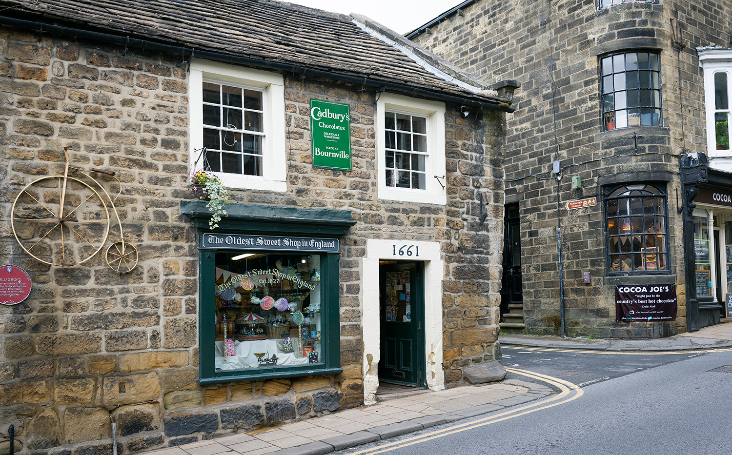 The exterior of the Oldest Sweet Shop is in an old stone building on a paved road and next to another old building.