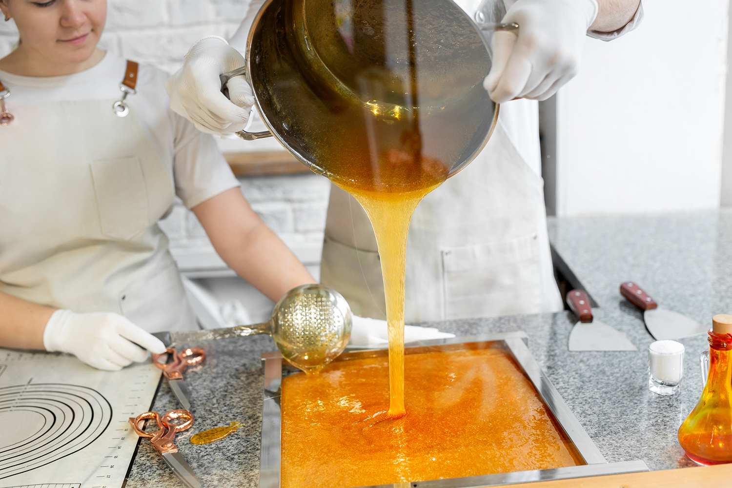 The gloved hands of a cook or chef are seen pouring caramelized sugar from a pot to a tray as another person observes.