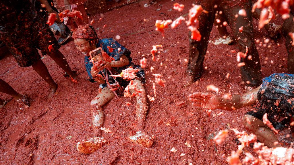 A participant covered in tomatoes and tomato juice sits on the tomato-covered ground and takes a selfie.