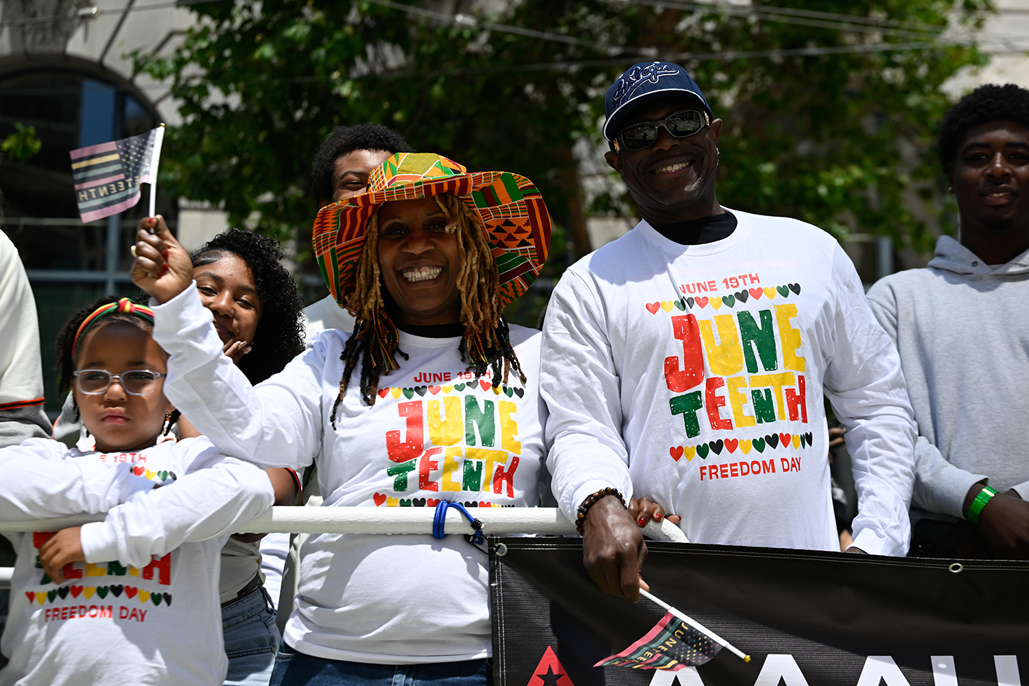 Smiling parade spectators wear Juneteenth t shirts and one woman holds a flag that says Juneteenth.