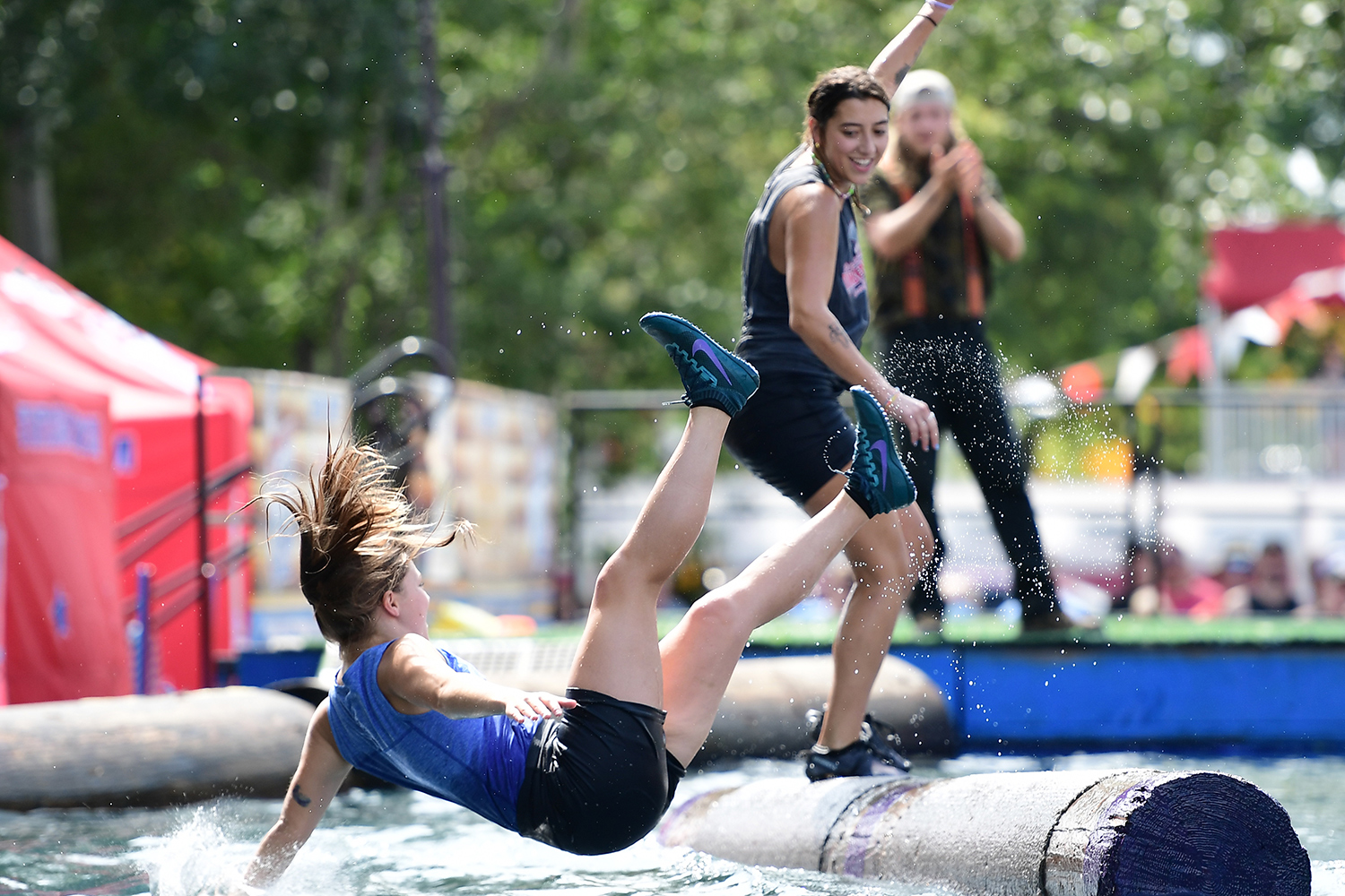 A woman balancing on a floating log watches as another woman falls off a log and into the water.