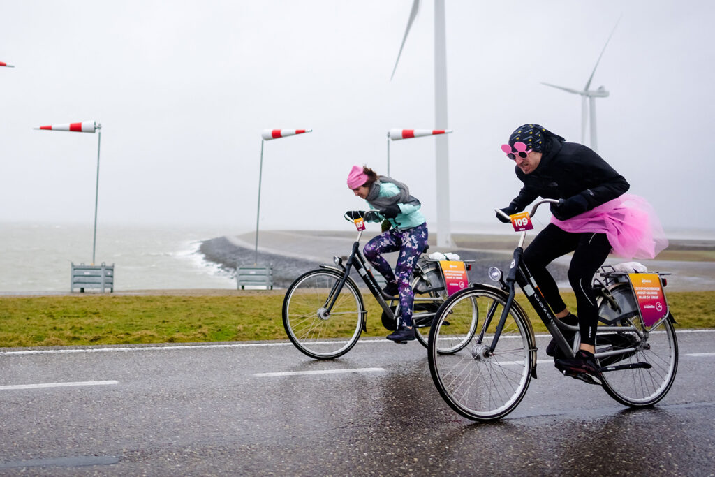 Two cyclists wearing colorful clothing race along a wet road next to a choppy body of water.