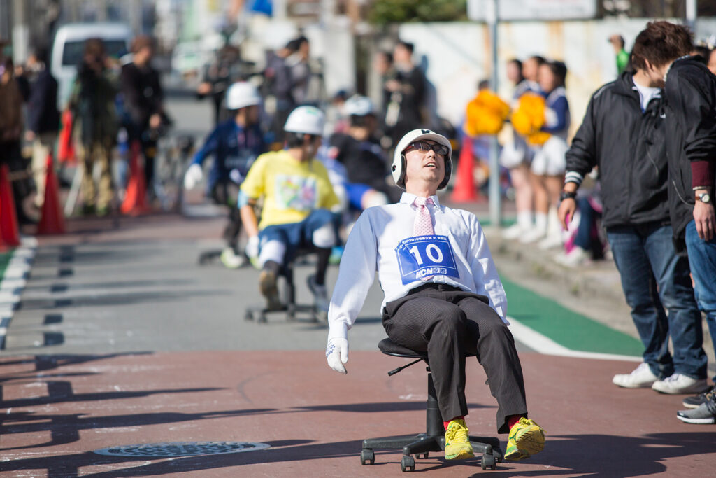 A man wearing a helmet and sitting on an office chair has his eyes closed as he and others take part in a chair race.