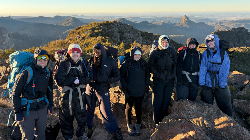 Seven teen girls pose from a high altitude with hills in the background.