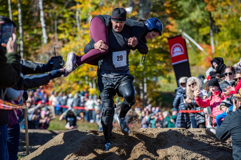 A man carries a woman who is draped across his shoulders as he move over a mount of dirt in front of spectators.