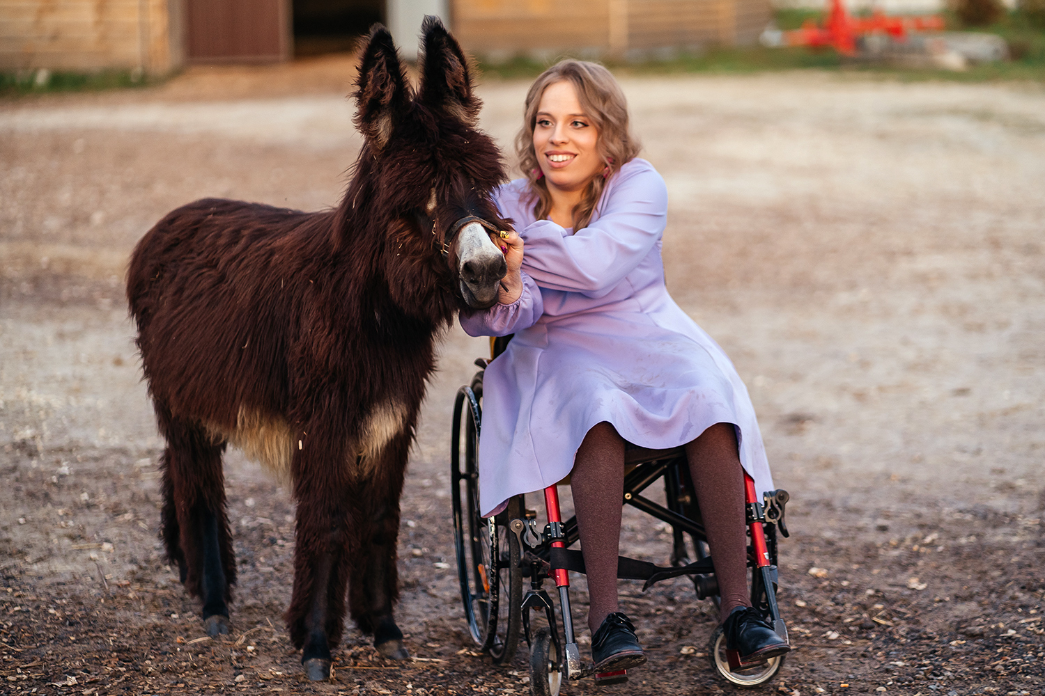 A woman who uses a wheelchair smiles and poses next to a shaggy brown donkey.