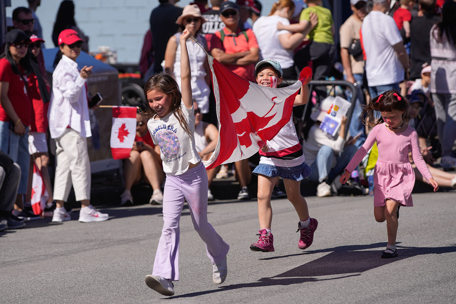 Three children walk down a blocked off street carrying a Canadian flag as spectators stand behind the barricades.