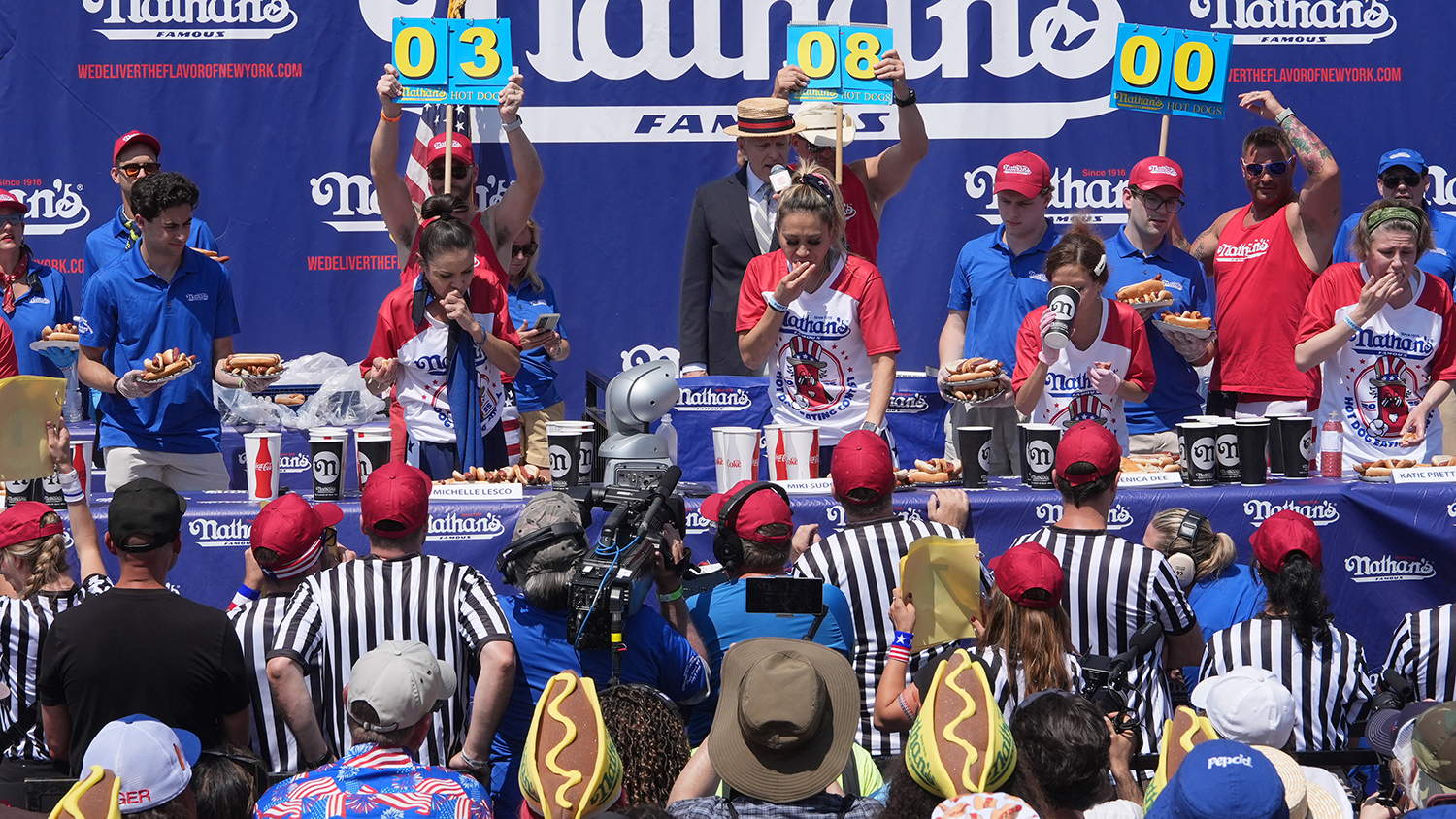 Four women stand at a table eating hot dogs as people behind them hold up numbers keeping count and an audience watches.
