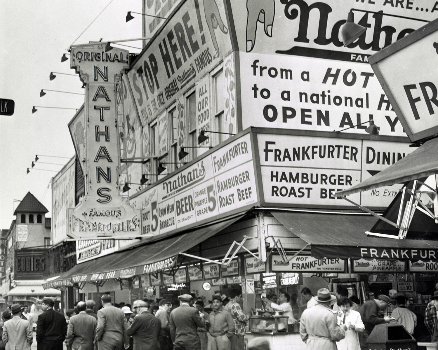 The Nathan’s Famous location on the boardwalk of Coney Island has multiple signs advertising its food and many people crowd and walk around it.