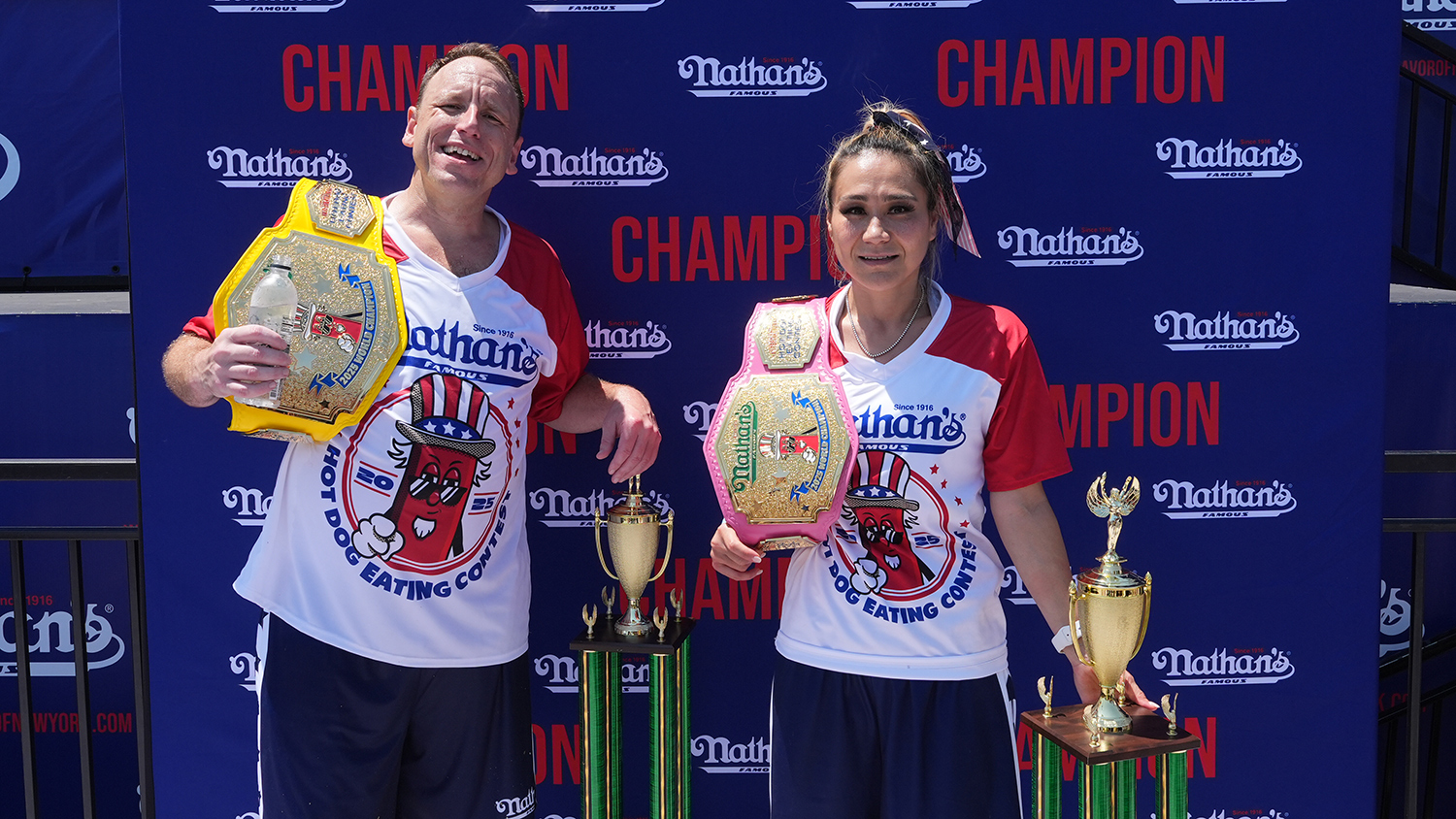 Joey Chestnut and Miki Sudo pose in front of a backdrop that says Nathan’s Champion with large trophies and have large gold winners’ belts slung on their shoulders.