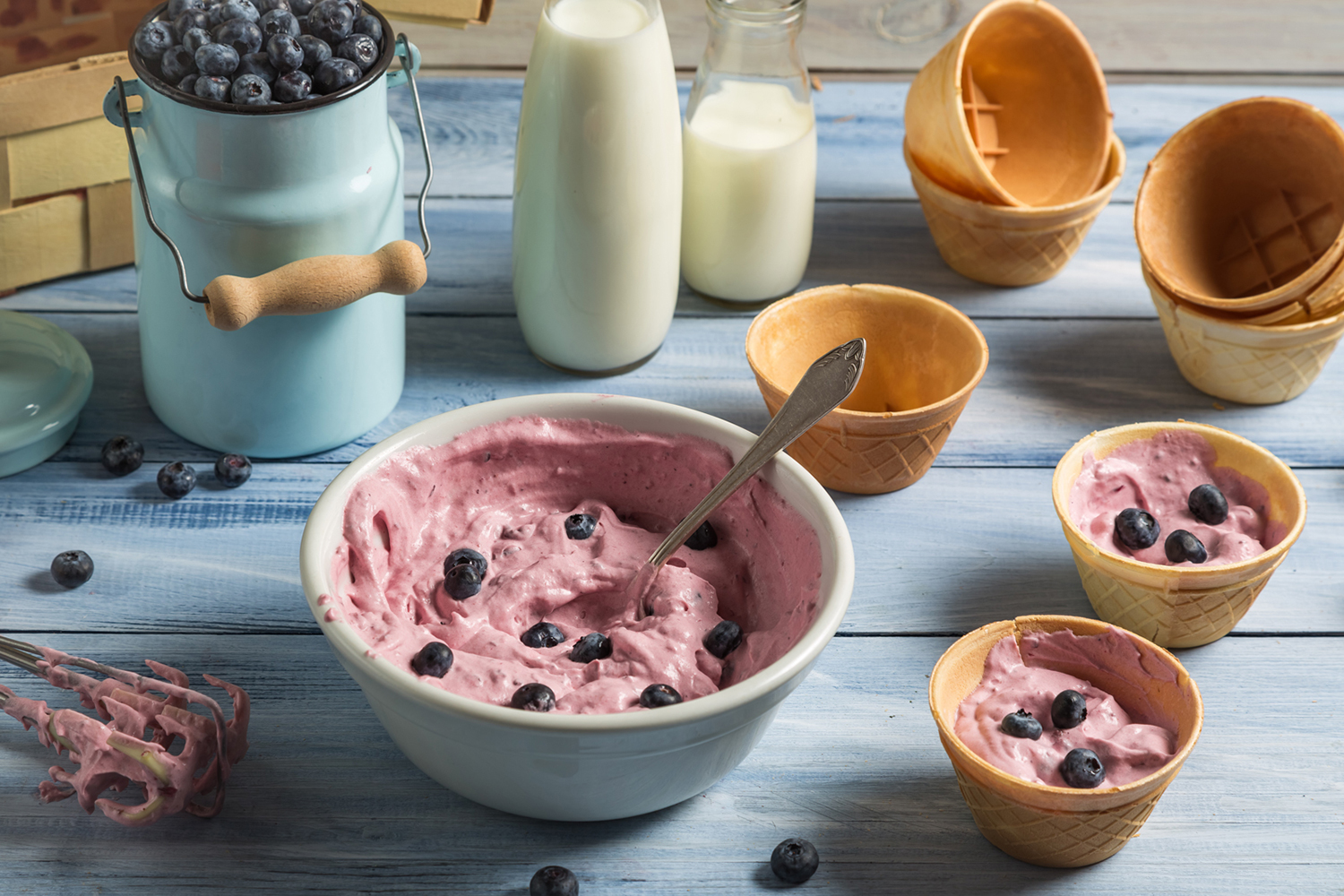 Blueberry ice cream is in a large bowl and two smaller bowls. Other bowls, bottles of milk, and blueberries also appear.