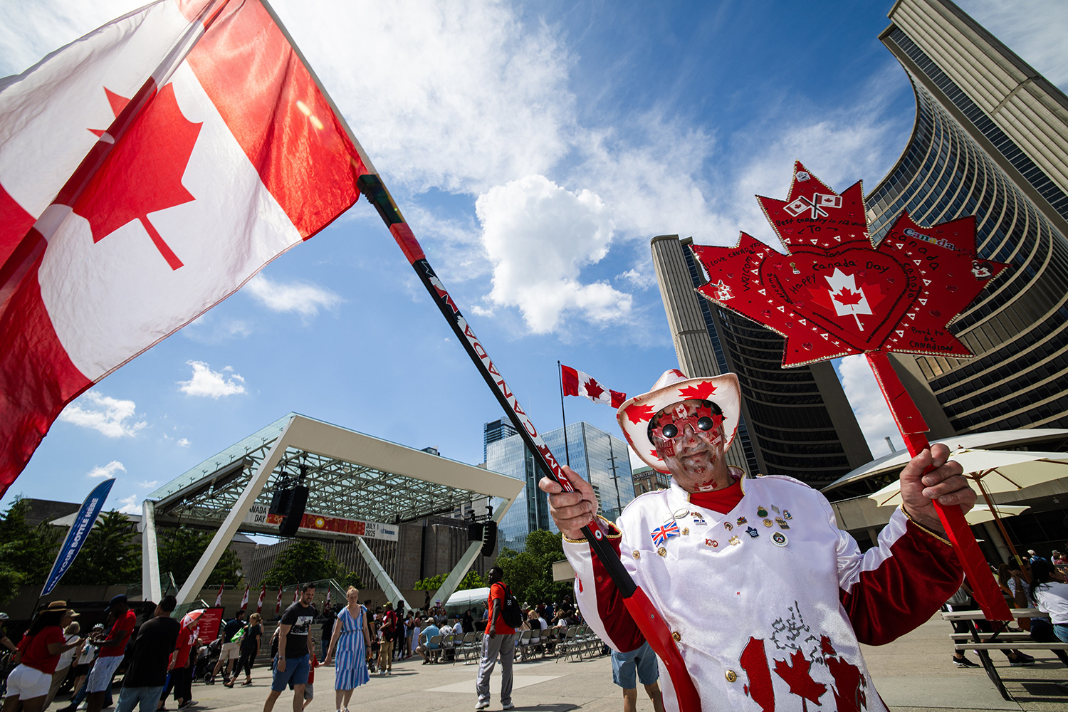 A man in a hat and an outfit with Canadian maple leaves holds a Canadian flag and a sign in the shape of a maple leaf.