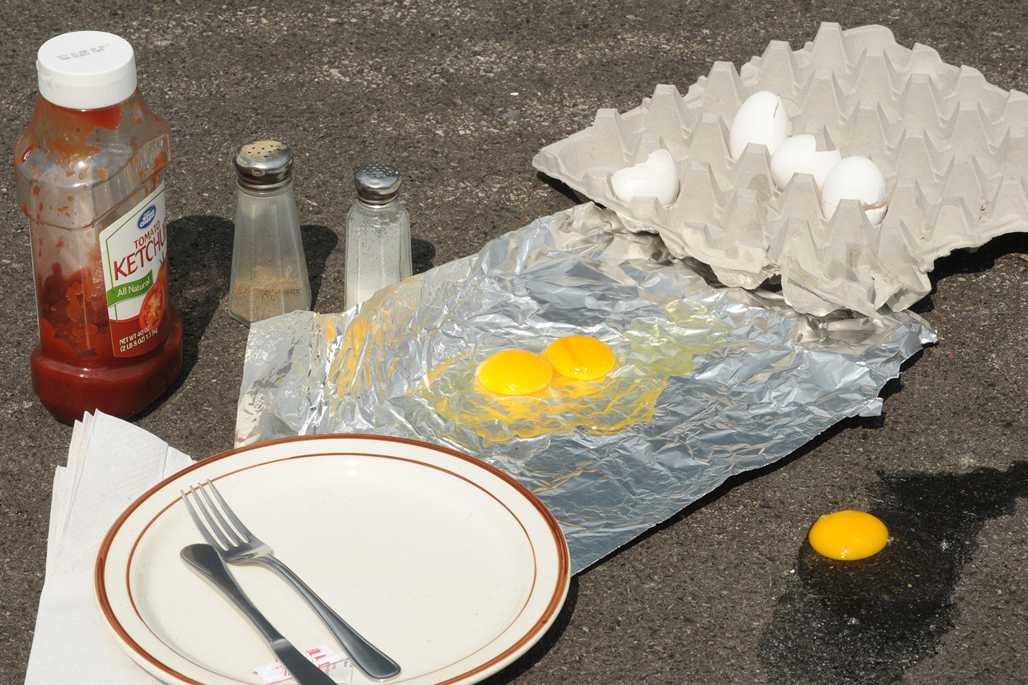Two raw eggs sit on aluminum foil that has been placed on an outdoor blacktop next to a partial carton of eggs, salt and pepper, ketchup, and a plate and silverware.
