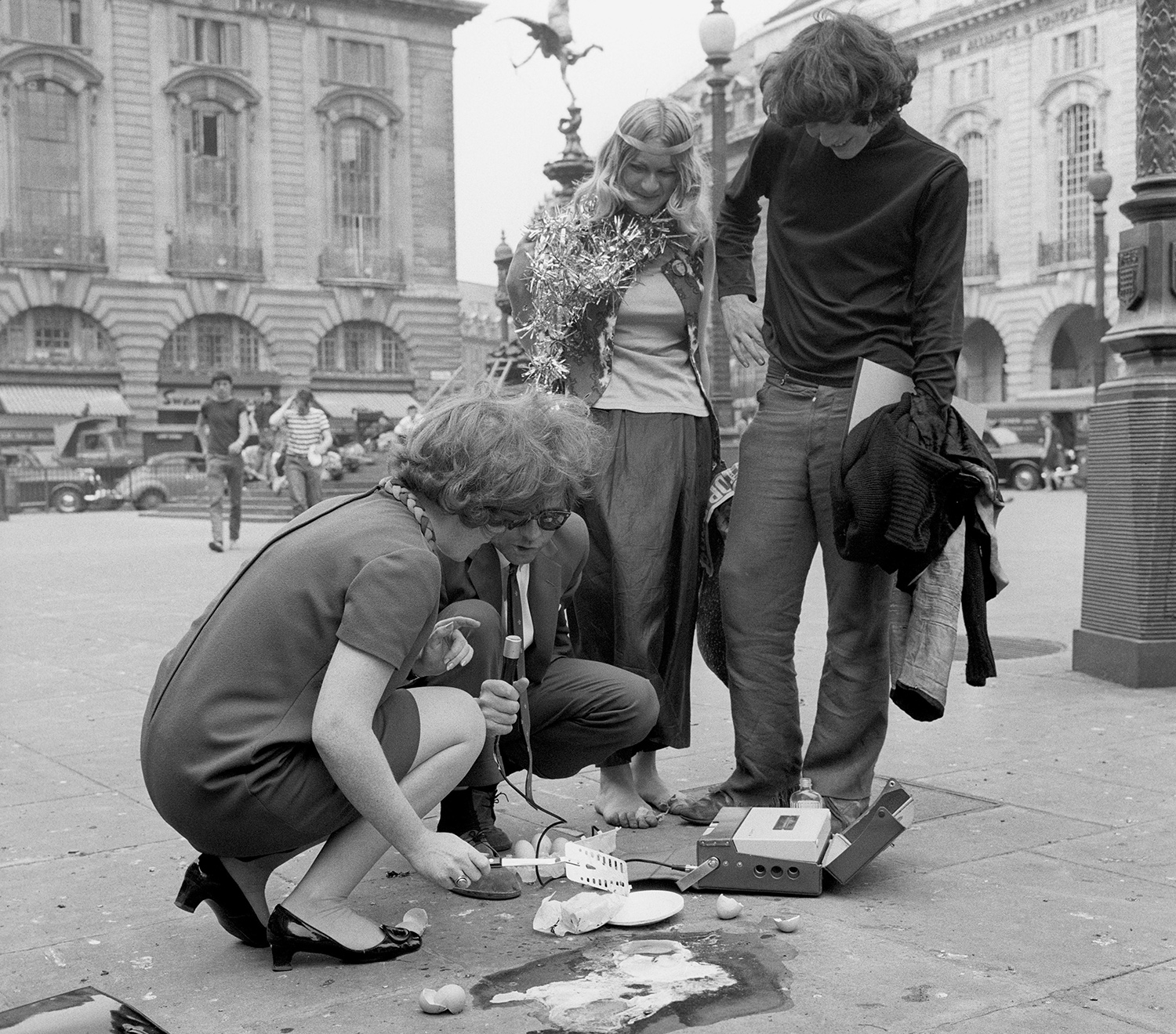 Two people attempt to fry an egg on a sidewalk and speak into radio equipment as two other people watch.