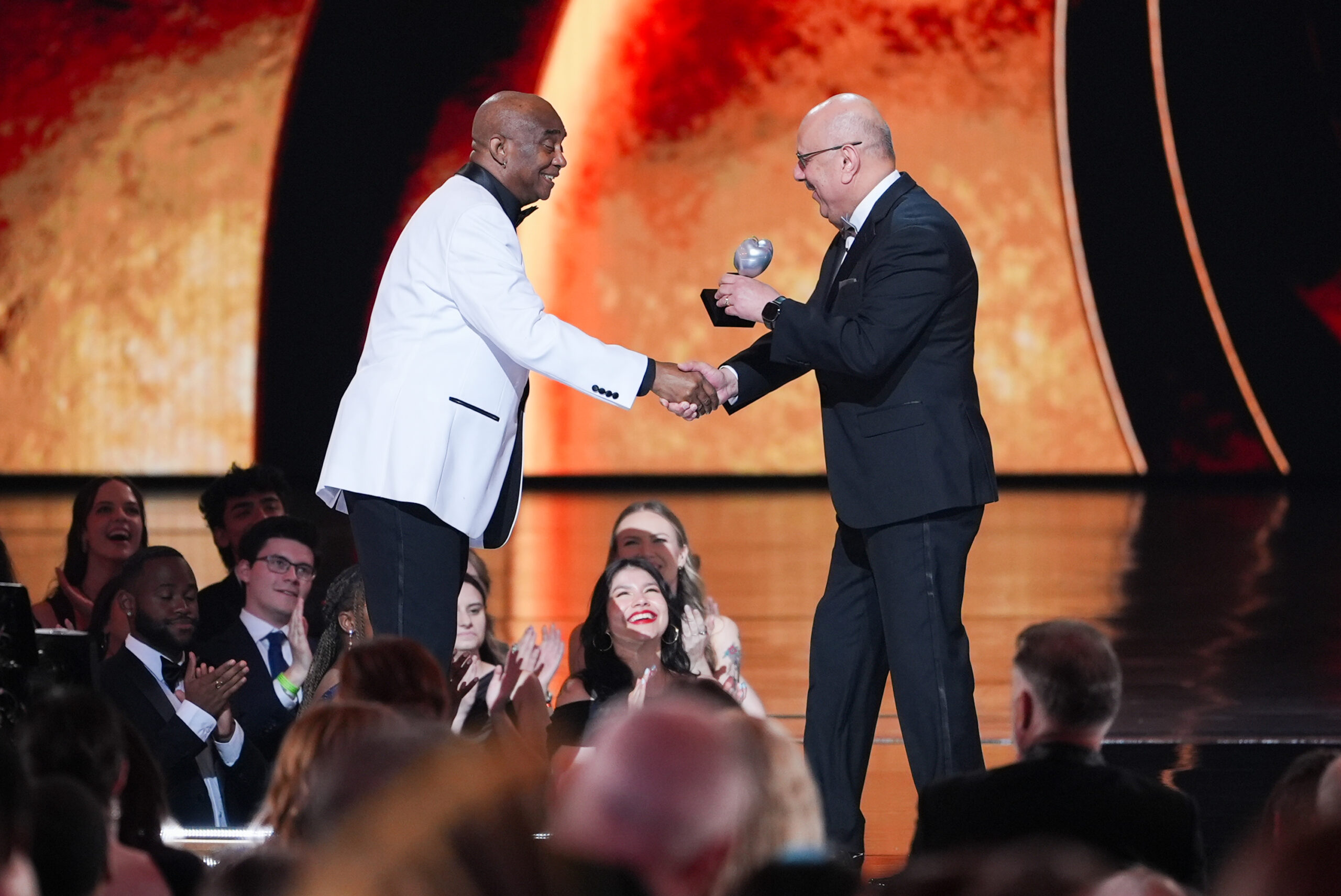 Freddie Hendricks stands on the stage and shakes the hand of, and accepts a Tony from, Farnam Jahanian, while audience members applaud.