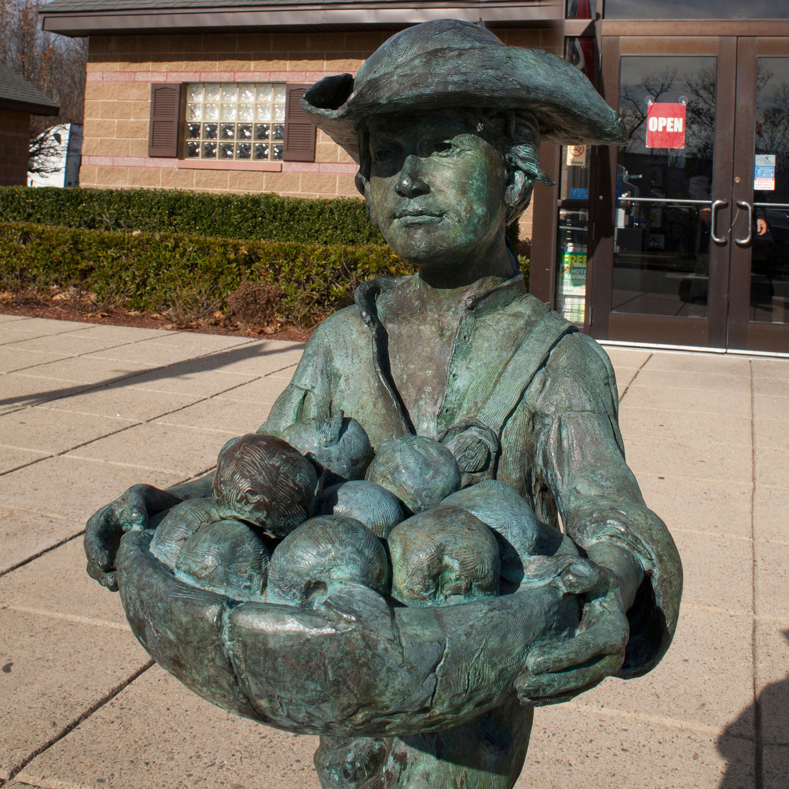 A metal statue of Johnny Appleseed wearing a hat and carrying a bowl of apples is in front of a building.