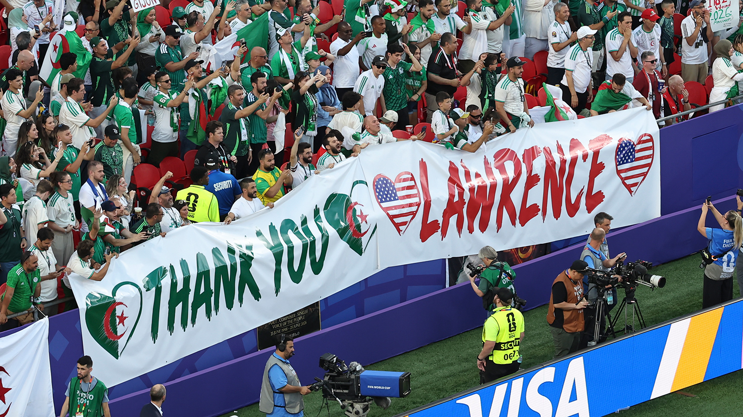People wearing green and white Algerian team jerseys are in the stands of a stadium displaying a large banner that says thank you Lawrence.