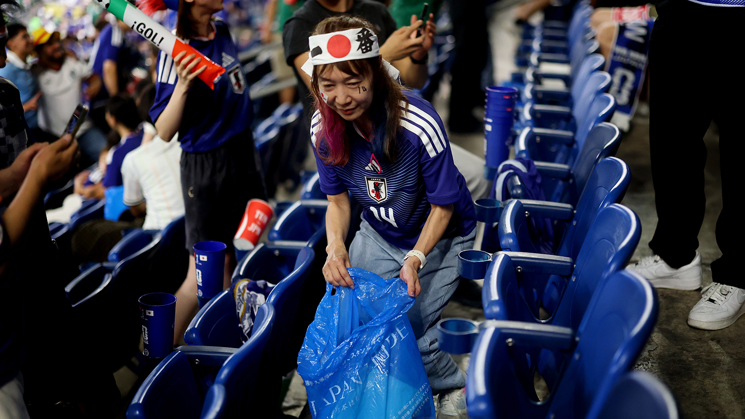 A fan wearing a Japanese team jersey is in the stands preparing to put garbage into a blue plastic bag.