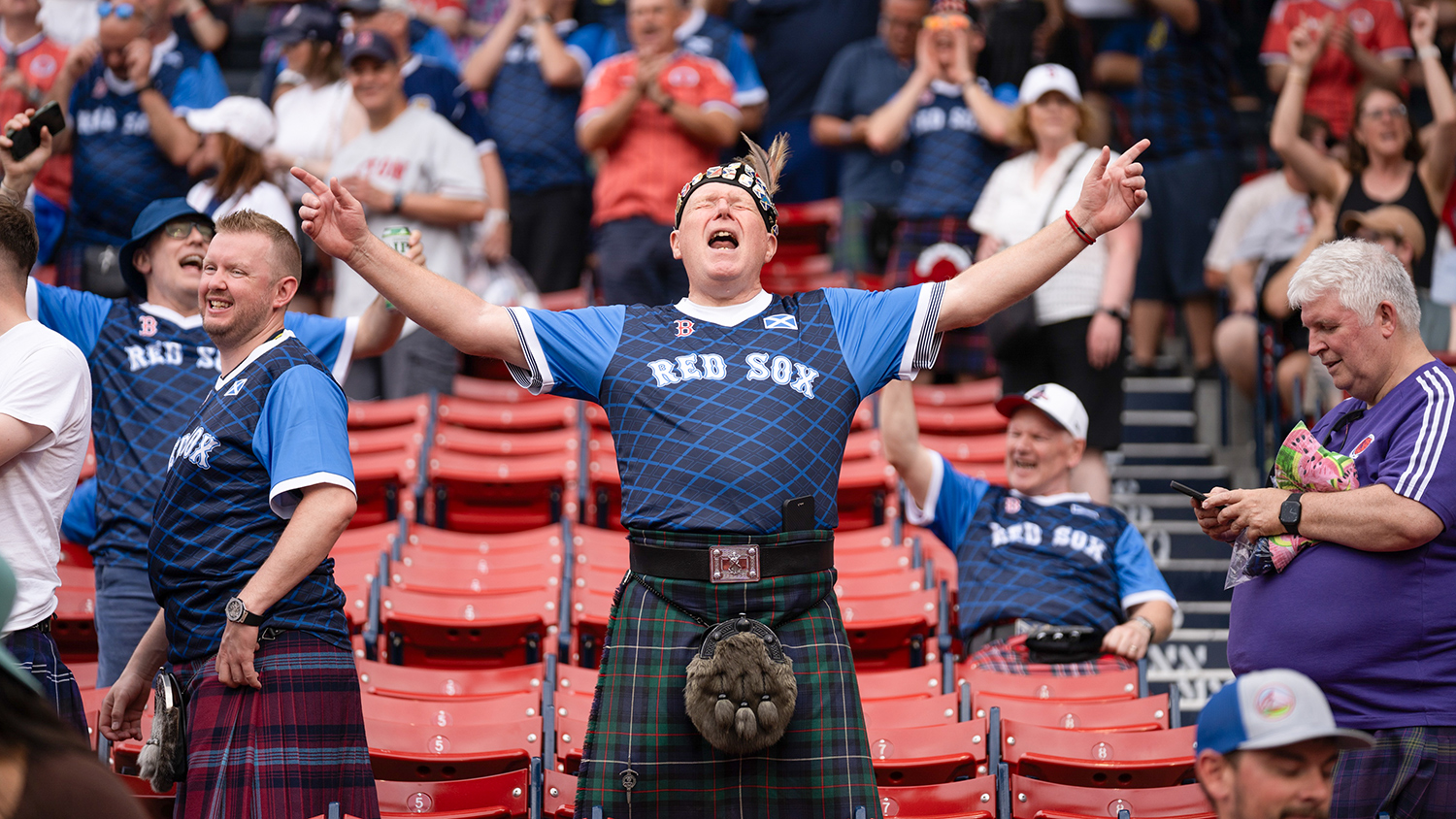 A fan in a Red Sox jersey and a kilt makes a joyful gesture as similarly dressed fans smile in the stands of Fenway Park.