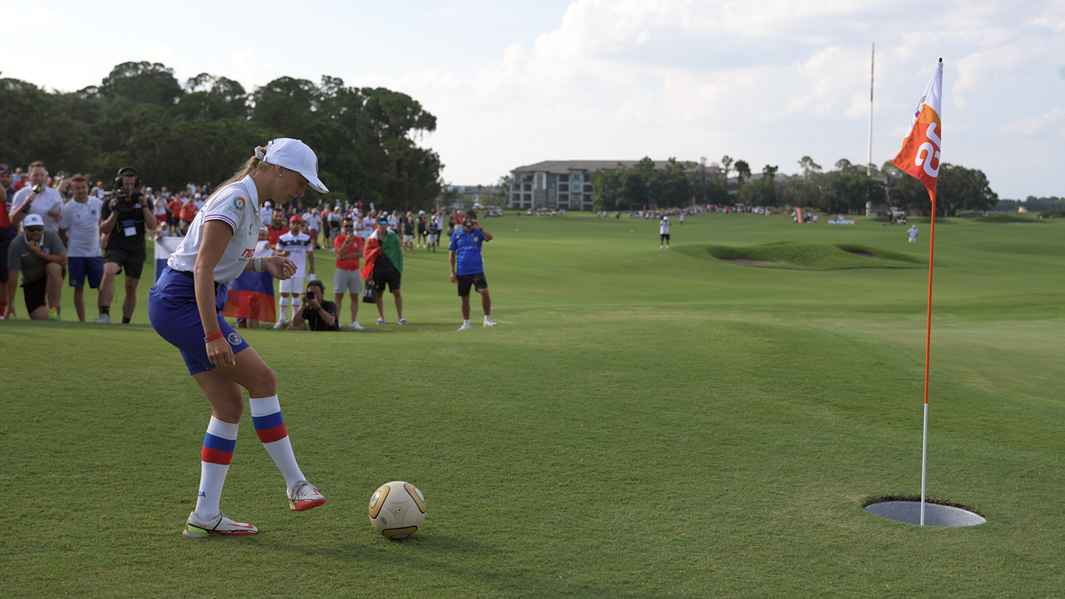 On a grassy course, Lucia Cermakova is about to kick a soccer ball toward a large hole with a flag in it.