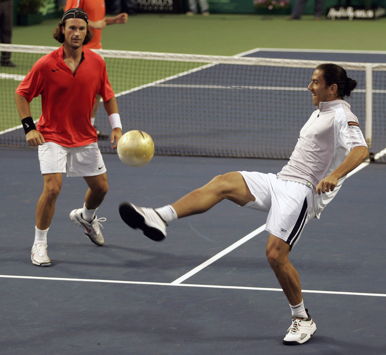 Two athletes stand on one side of a tennis net, and one of them is about to kick a soccer ball.