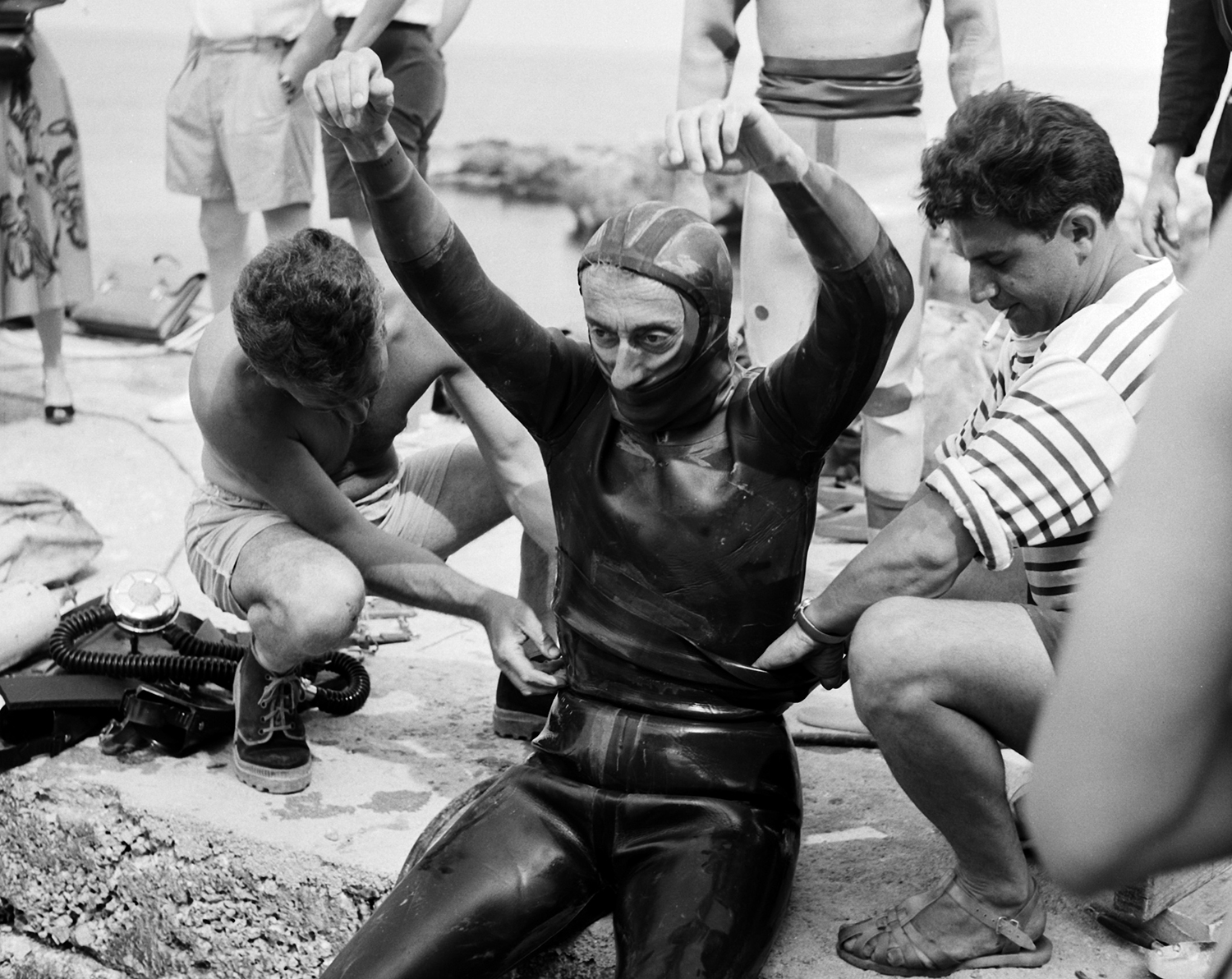 Jacques Cousteau sits on a sea wall with his arms up as two men help him finish putting on his diving gear.