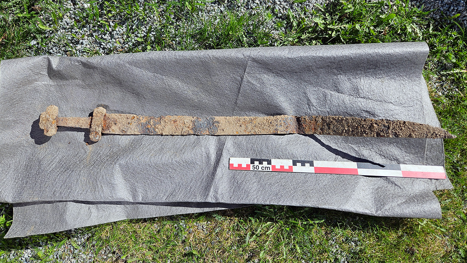 A rusty metal Viking sword is lying on a piece of cloth and is about twice as long as a 50-centimeter measuring tape.