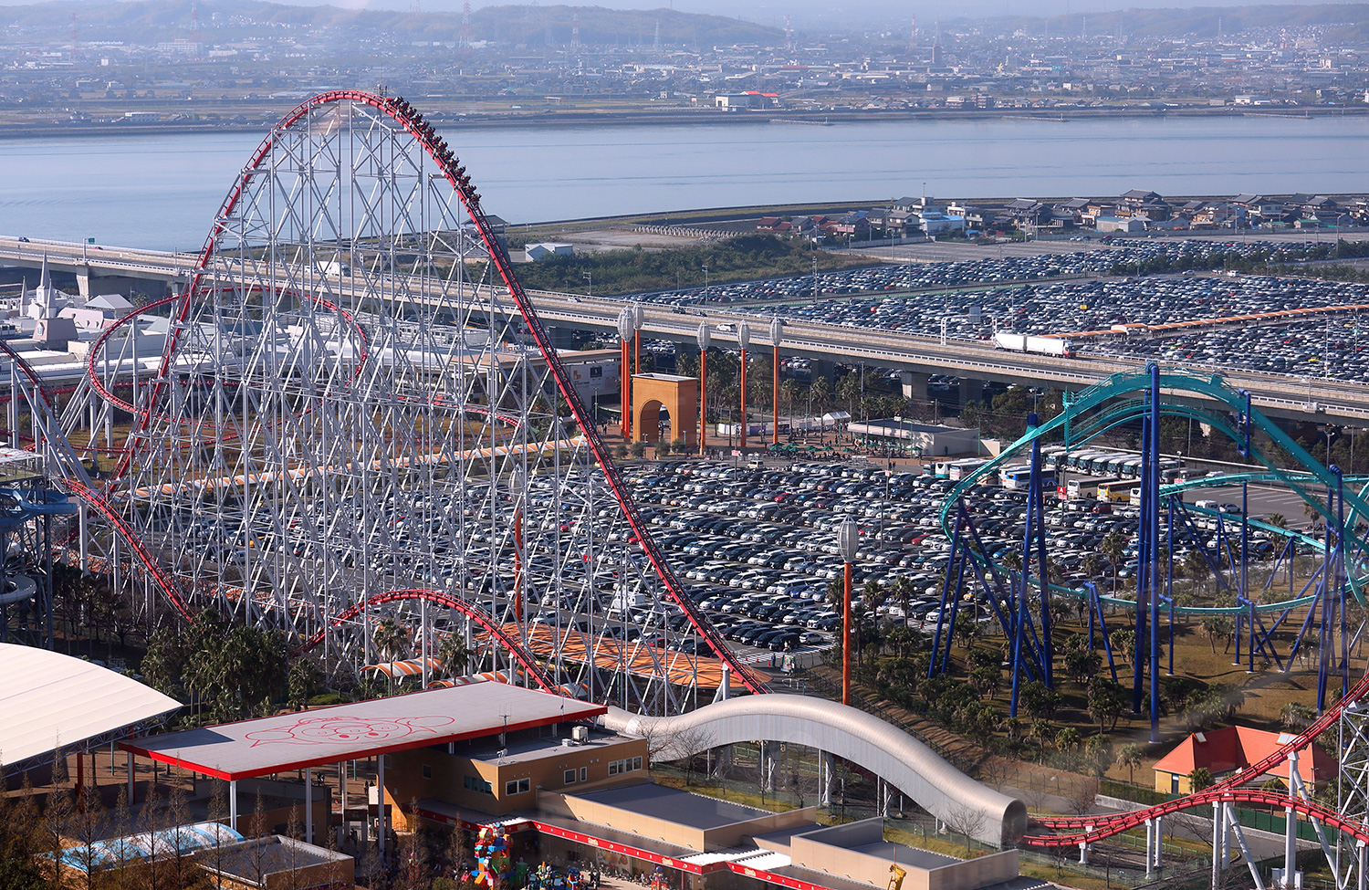 Side shot of a large roller coaster among other rides and a parking lot.