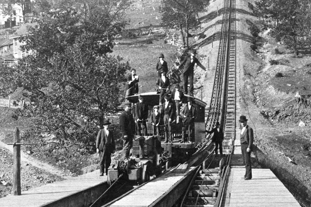 Two sets of tracks have been constructed on a steep hill. Men in 1800s clothing stand at the bottom next to a car that is on the tracks.