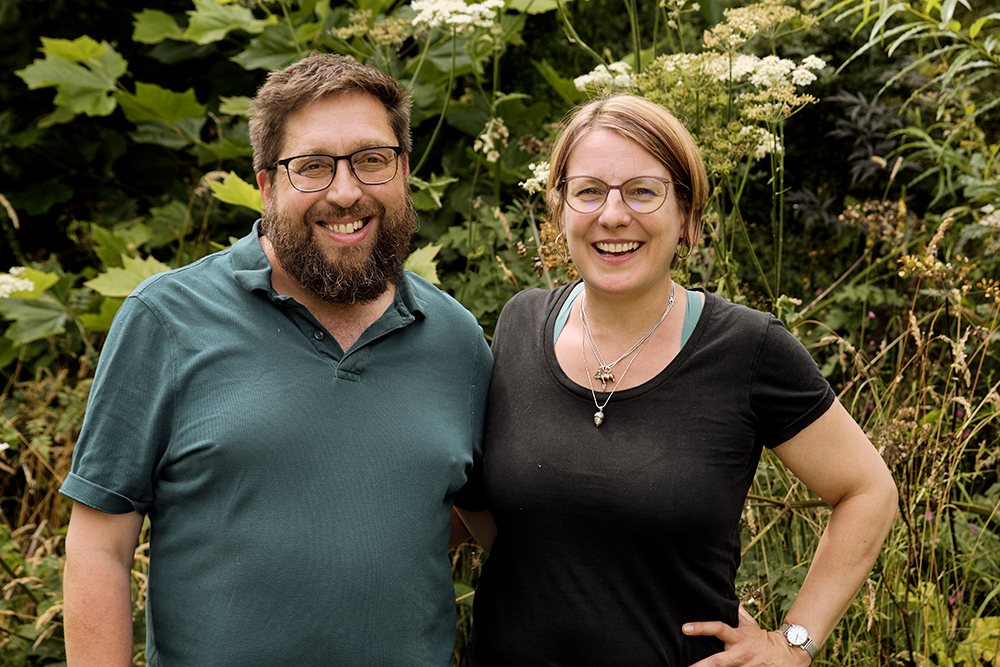 A smiling Gavin and Alice Munro pose together in front of outdoor vegetation.