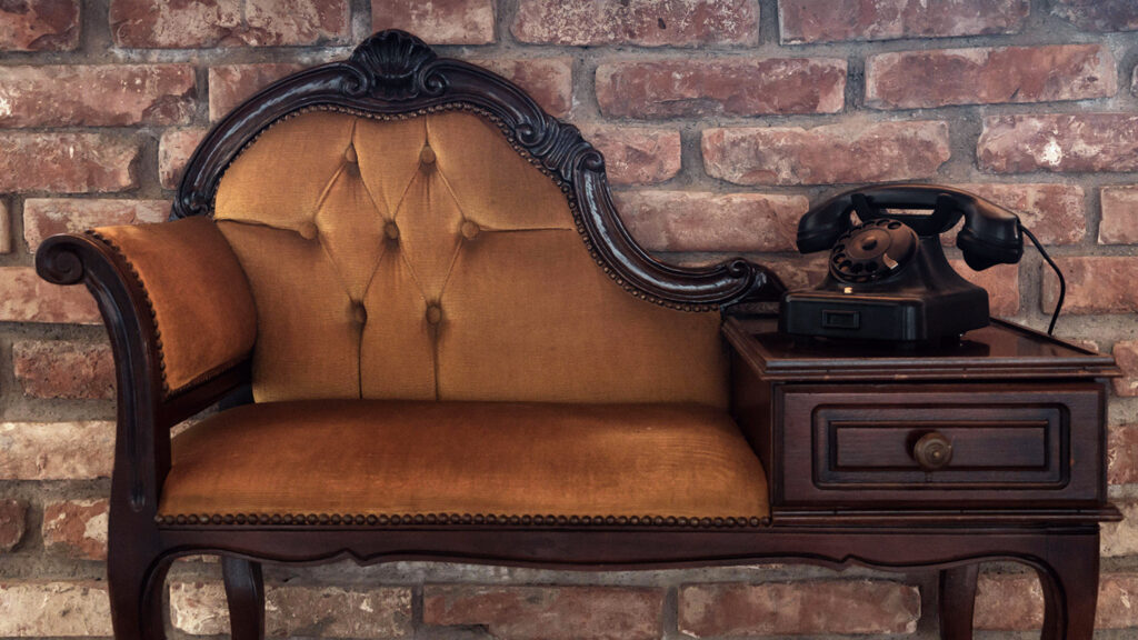 A piece of Victorian furniture in the shape of a settee consists of a plush seat on one end and a table with a telephone on the other.