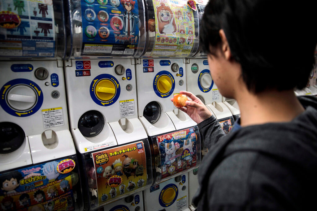 A child takes a capsule out of a vending machine that sells toys or stickers and is next to similar vending machines.
