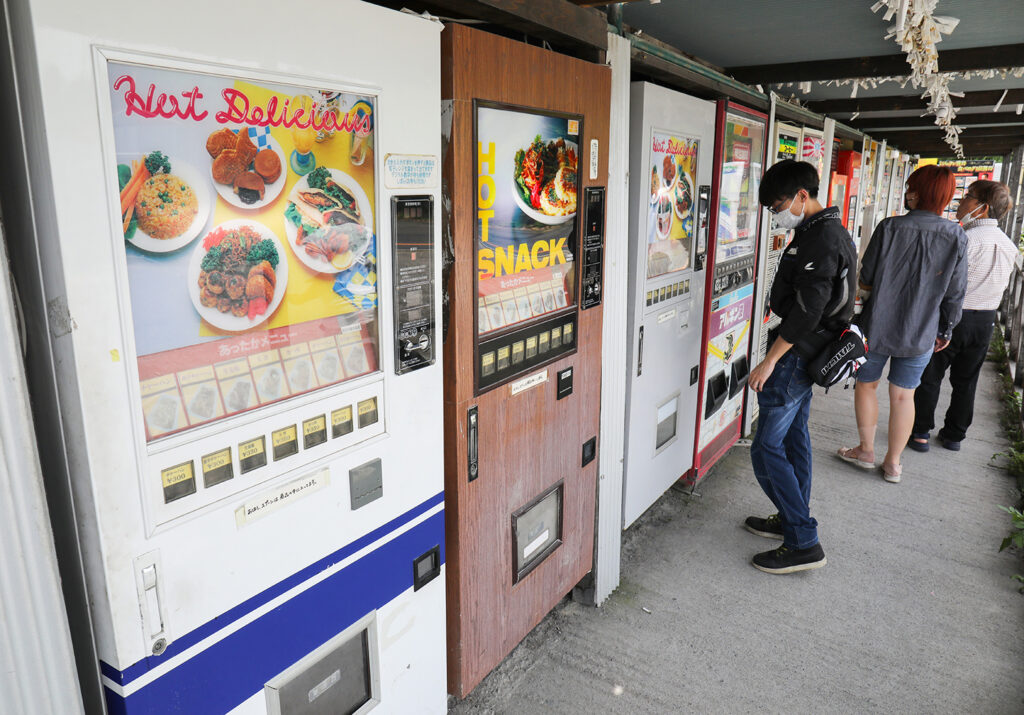 Three people look at the selections in some food vending machines that are among many in a long row.