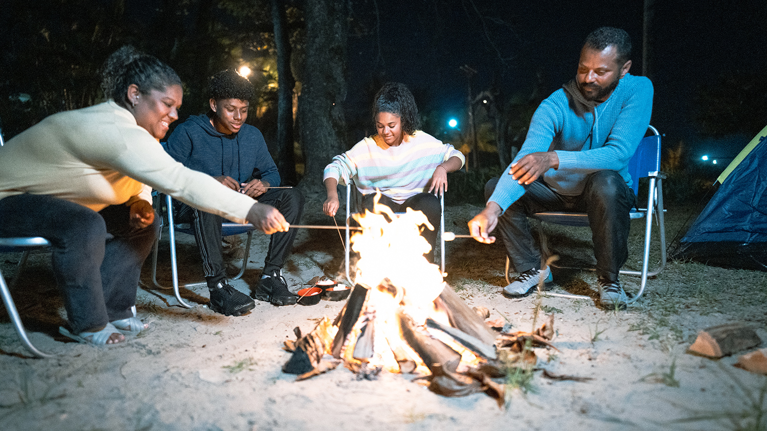 A family of four sits on a sandy beach and roasts marshmallows over a campfire.
