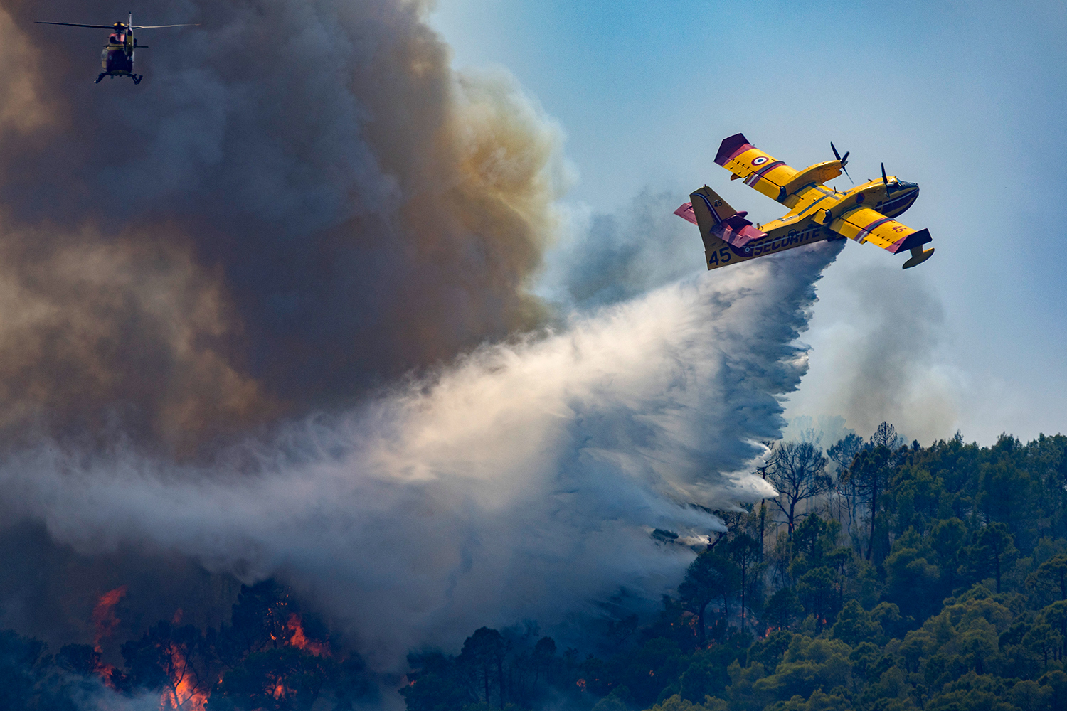 A yellow biplane drops a firefighting substance over a forest that is burning.
